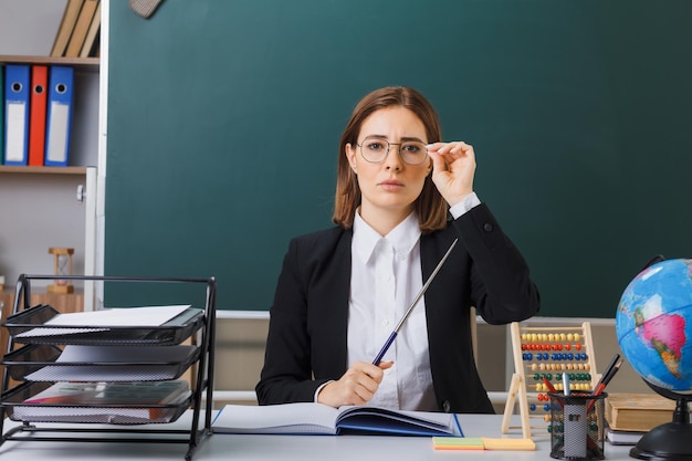 Young woman teacher wearing glasses sitting at school desk in front of blackboard in classroom with abacus and globe checking class register holding pointer looking confident