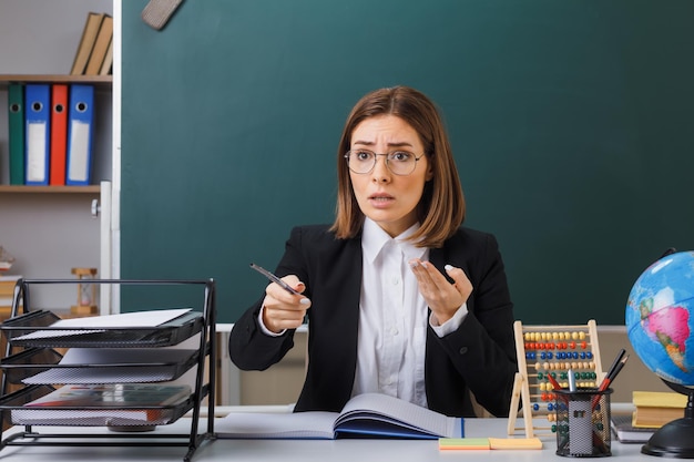 Young woman teacher wearing glasses sitting at school desk in front of blackboard in classroom with abacus and globe checking class register holding pointer raising arm in displeasure confused