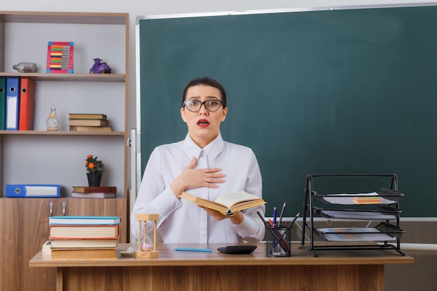 Young woman teacher wearing glasses sitting at school desk with book in front of blackboard in classroom looking at camera being worried