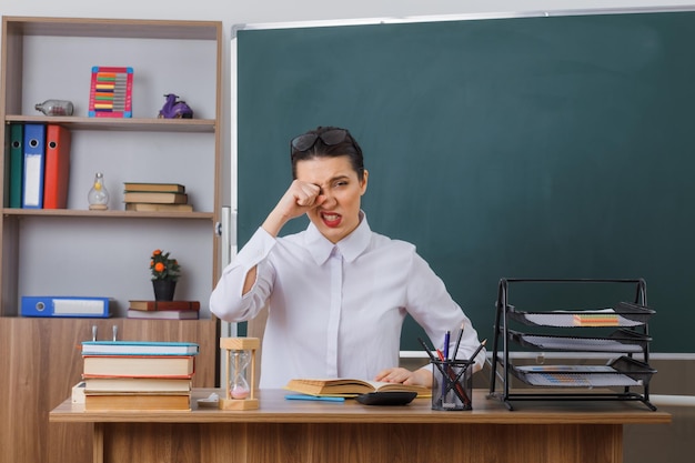 Young woman teacher wearing glasses sitting at school desk with book in front of blackboard in classroom looking tired rubbing eye with fist