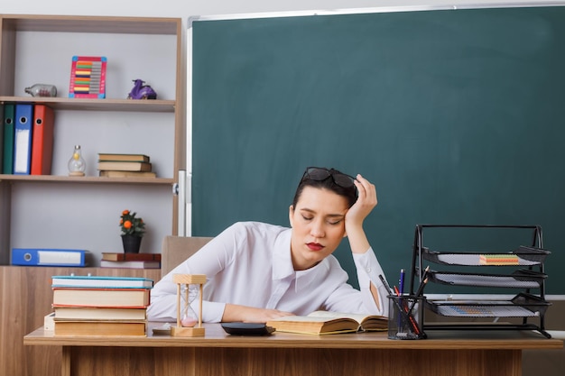 Young woman teacher wearing glasses sitting at school desk with book in front of blackboard in classroom looking tired sleeping leaning head on her palm