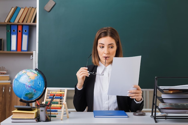 Insegnante di giovane donna con gli occhiali seduto al banco della scuola con il globo e i libri davanti alla lavagna in classe che tiene il foglio di carta vuoto bianco che sembra incuriosito