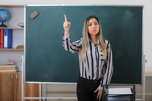 young woman teacherstanding near blackboard in classroom explaining lesson showing index finger with serious face