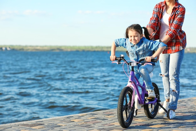 Young woman teaching her daughter to ride bicycle outdoors near river