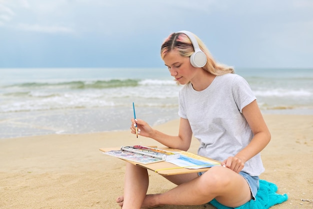 写真 海のビーチに座っている水彩画でスケッチを描くヘッドフォンで若い女性のティーンエイジャー