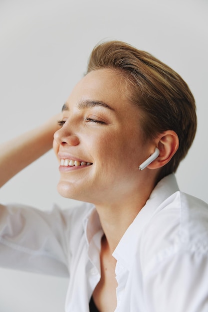 Young woman teenager listening to music with infertile headphones and dancing home grinning with teeth with a short haircut in a white shirt on a white background Girl natural poses with no filters