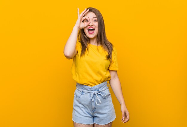 Young woman teenager wearing a yellow shirt excited keeping ok gesture on eye.