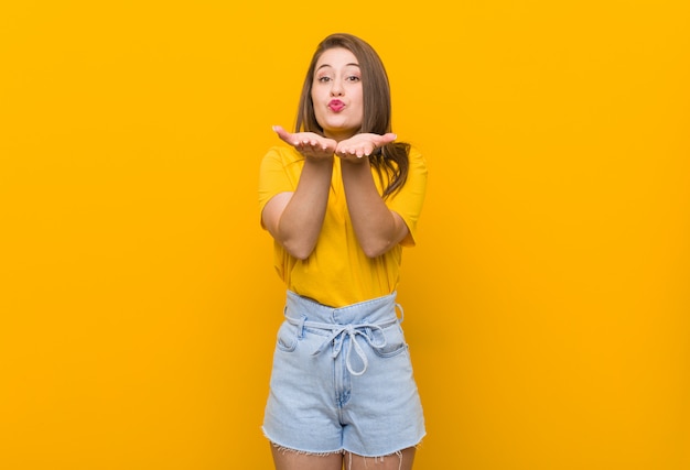 Young woman teenager wearing a yellow shirt folding lips and holding palms to send air kiss.