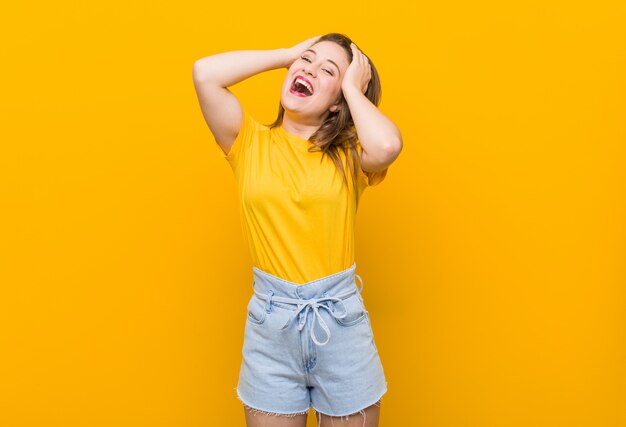 Young woman teenager wearing a yellow shirt laughs joyfully keeping hands on head. Happiness concept.