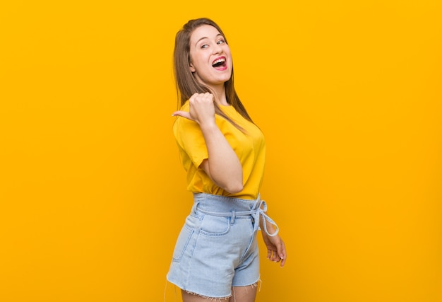 Young woman teenager wearing a yellow shirt points with thumb finger away, laughing and carefree.