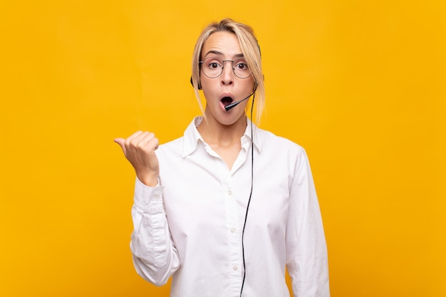 Young woman telemarketer looking astonished in disbelief, pointing at object on the side