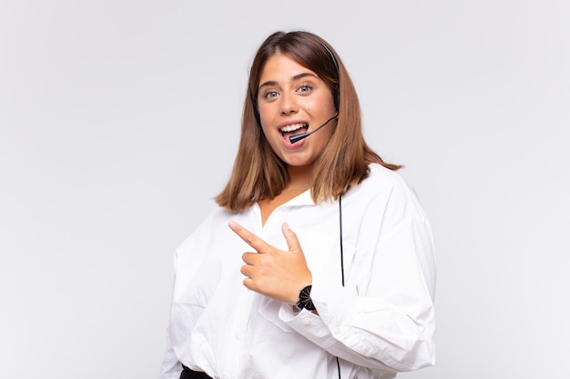 Young woman telemarketer looking excited and surprised pointing to the side and upwards to copy space