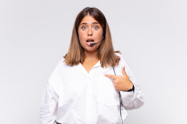 Young woman telemarketer looking shocked and surprised with mouth wide open, pointing to self