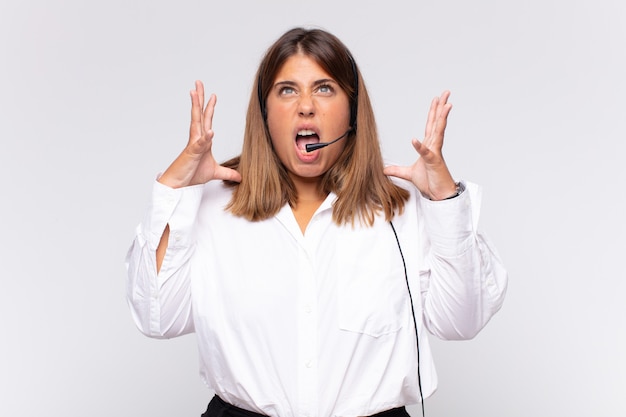 Young woman telemarketer screaming with hands up in the air, feeling furious, frustrated, stressed and upset