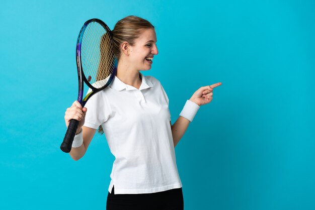 Young woman tennis player isolated on blue background pointing finger to the side and presenting a product