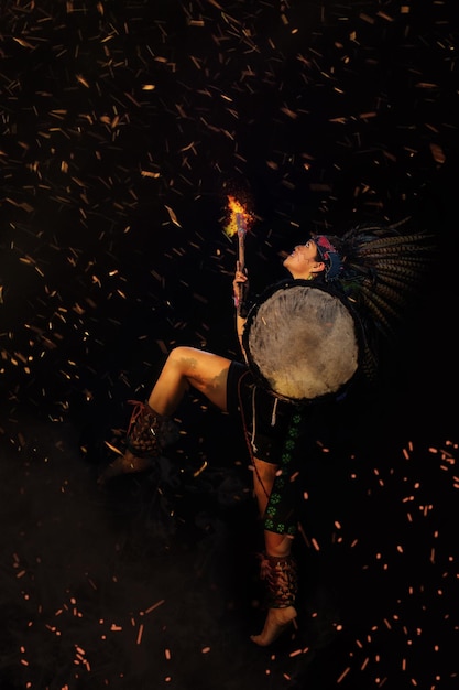 Молодая женщина Теотиуакана, Ксикаланка - тольтеков на черном фоне, с традиционным танцевальным платьем с атрибутами с перьями и барабаном, свет над головой, фон в огне