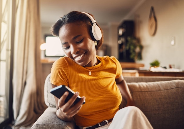 젊은 여성이 전화로 소셜 미디어를 탐색하고 스크롤하면서 행복하고 평온한 기분을 느끼고 소파에서 휴식을 취하면서 음악 팟캐스트를 듣거나 재미있는 인터넷 밈을 온라인으로 시청합니다.