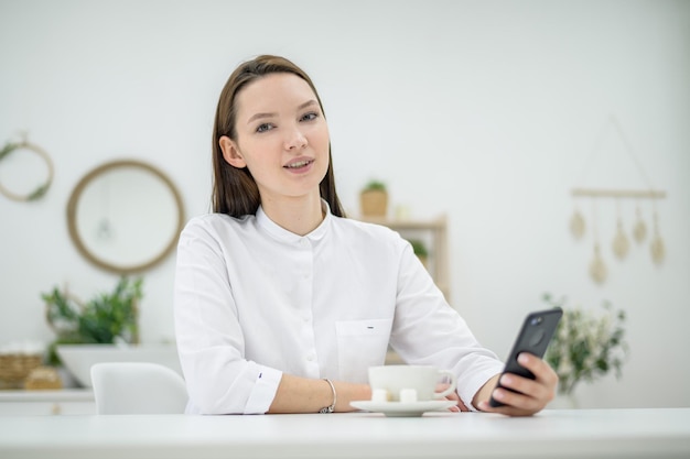 仕事の休憩中に電話でテキスト メッセージを送信する若い女性 女の子はオフィスでコーヒーを飲んでいます オフィス マネージャー