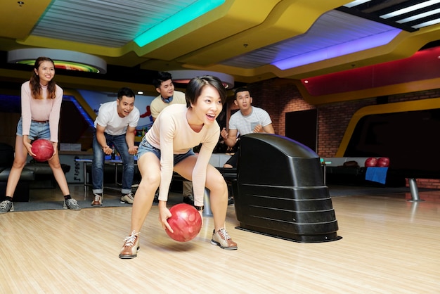 Young woman throwing ball while playing bowling together with her friends