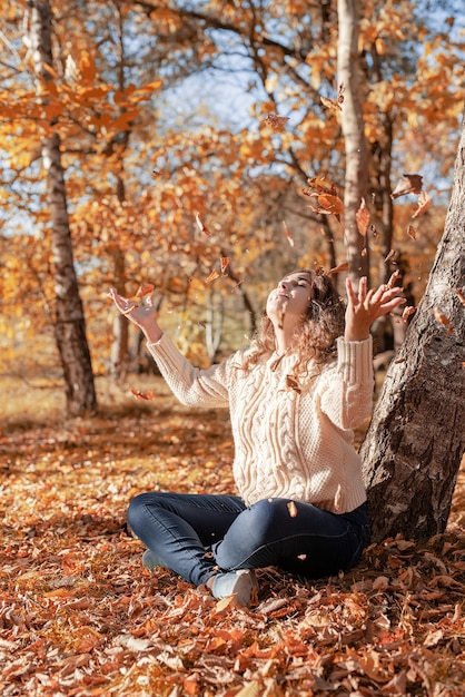 Foto giovane donna che lancia foglie gialle nell'aria seduta nella foresta autunnale