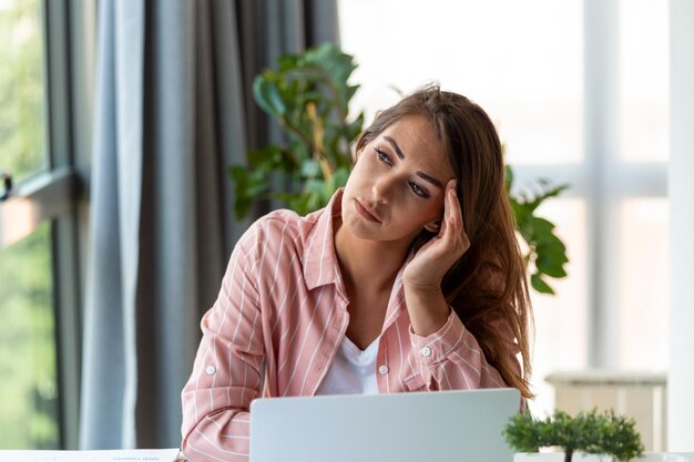 Photo young woman tired exhausted from working studying hard bored and frustrated looking at laptop head resting on hand bright space big windows at home concept stress concept