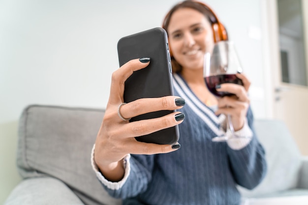 A young woman toasts with friends over a video call with red wine from the sofa