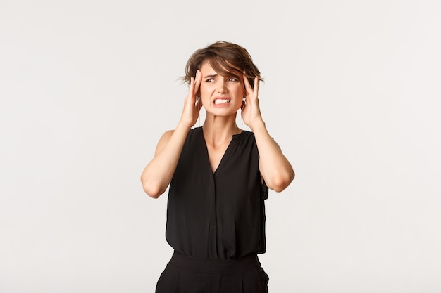 Young woman touching temples and grimacing from painful migraine. Girl complaining on headache, standing white.