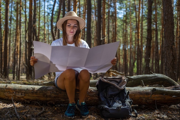 Turista della giovane donna in un cappello e una maglietta si siede su un tronco e guarda una mappa durante una sosta nella foresta.