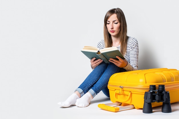 Young woman tourist is reading a book with a suitcase, binoculars on a light background