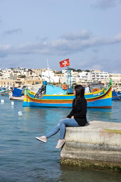 몰타의 Marsaxlokk에서 다채로운 보트로 가득한 바다를 배경으로 앉아 있는 젊은 여성 관광객