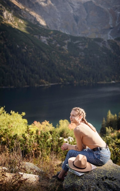 Молодая женщина-туристка сидит на горной вершине и в красивых горах. Пейзаж со спортивной девушкой, зеленый лес, холмы, небо, озеро. Путешествие и туризм.