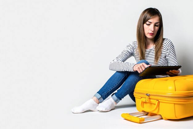 Young woman tourist sitting with a tablet near a yellow suitcase on a light background