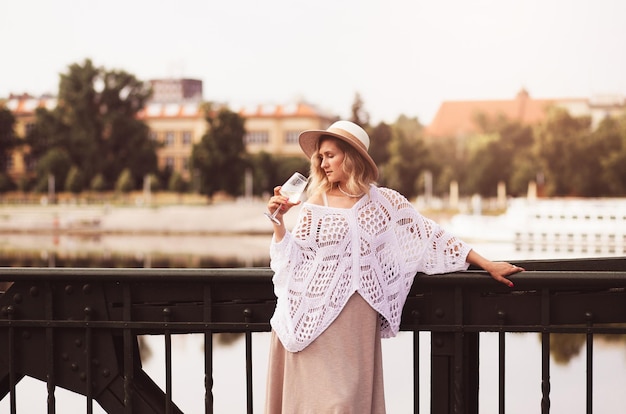 Turista della giovane donna in piedi con un bicchiere di vino bianco al ponte nel fiume della città europea sullo sfondo