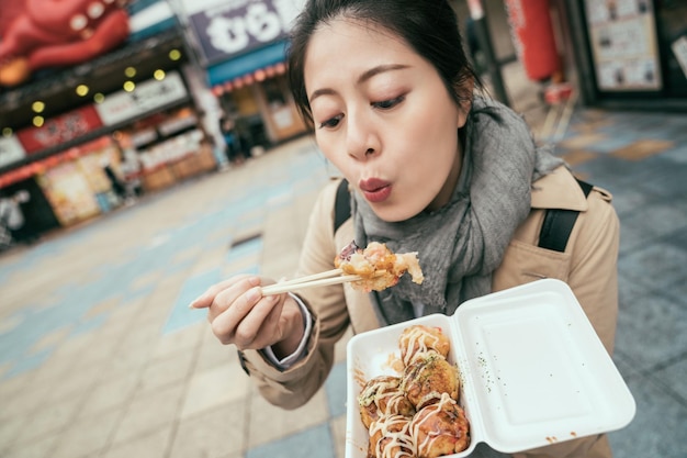 배경이 흐릿한 거리에서 야외에서 일본 현지 맛있는 음식을 맛보는 젊은 여성 관광객. 한국 여자 여행자는 상자를 들고 젓가락으로 뜨거운 타코야키를 불고 있습니다. 여자는 문어 공을 먹는다