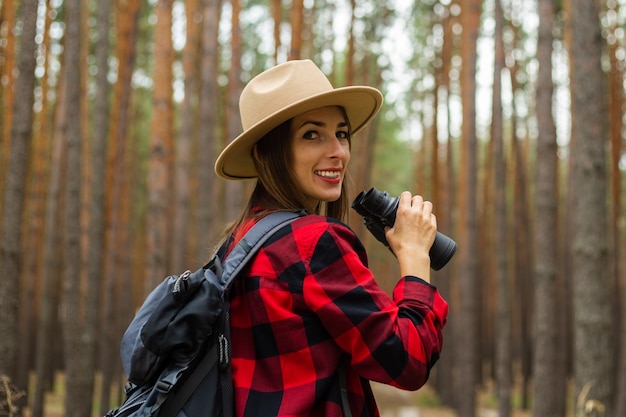 Young woman tourist with backpack, hat and red plaid shirt holding binoculars in the forest.