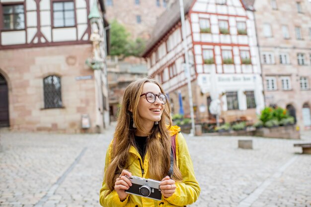 ドイツ、ニュルンベルク市の背景に美しい建物と古い広場にバックパックで立っている黄色のレインコートで若い女性観光客