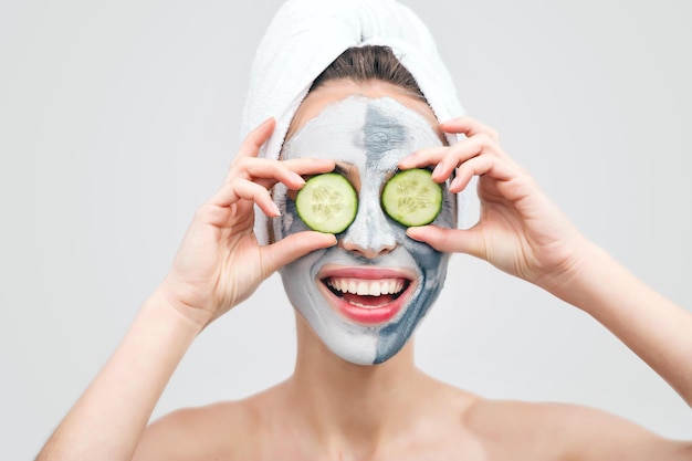 Young woman in towel with clay mask holding slices of cucumber against her eyes