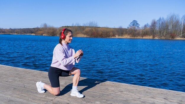 トラックスーツを着た若い女性が音楽を聴き、屋外でトレーニングをしている背景に澄んだ水のある池公園の新鮮な空気