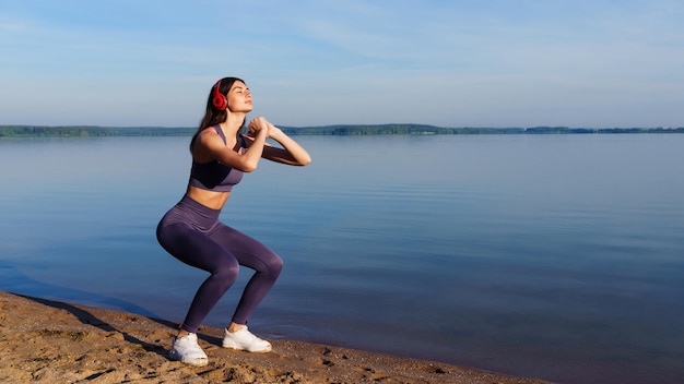 A young woman in a tracksuit squats during a workout outside in the morning in the summer