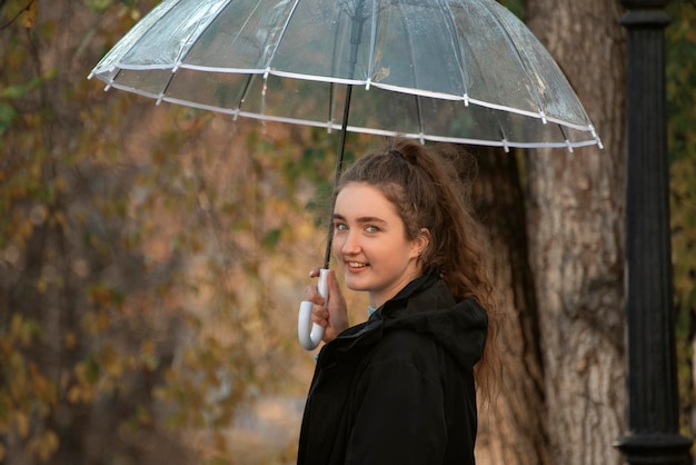 Young woman under transparent umbrella stands half turn to lens Girl with long hair in ponytail walks in autumn park