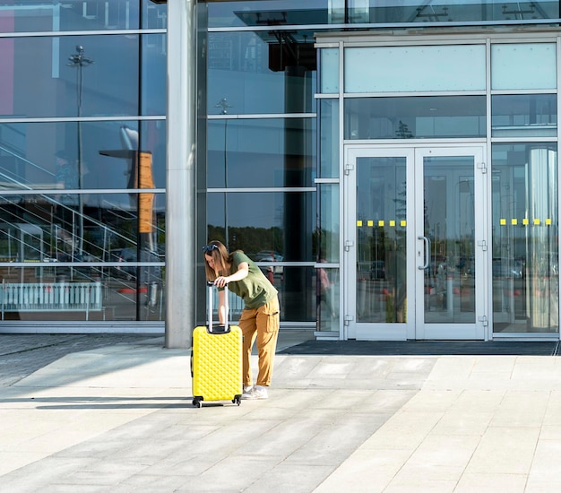 空港への入り口の横に黄色いスーツケースを持ってカジュアルな服を着た若い女性旅行者は、航空便の旅行と休暇の観光コンセプトの外にあります
