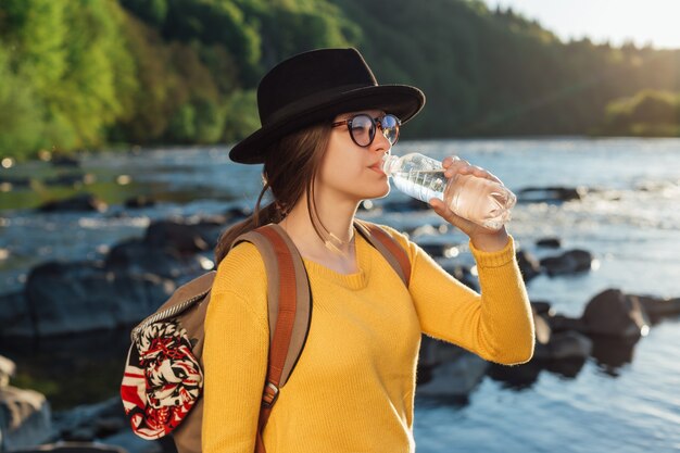 自然の川の背景にボトルから純粋な水を飲む若い女性旅行者