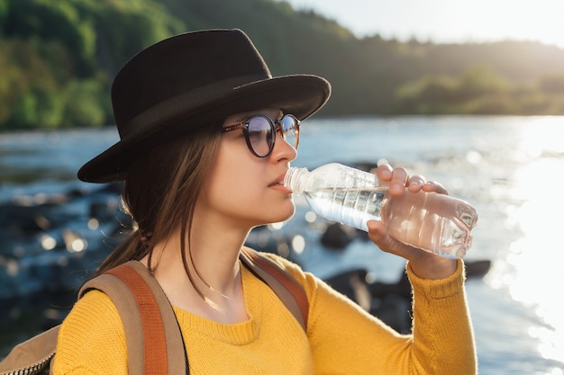 Young woman traveler drinking pure water from bottle on nature river background