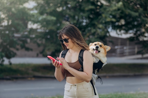 Young woman traveler is walking outdoors and using phone in\
city parkland with dog welsh corgi pembroke in a special\
backpack