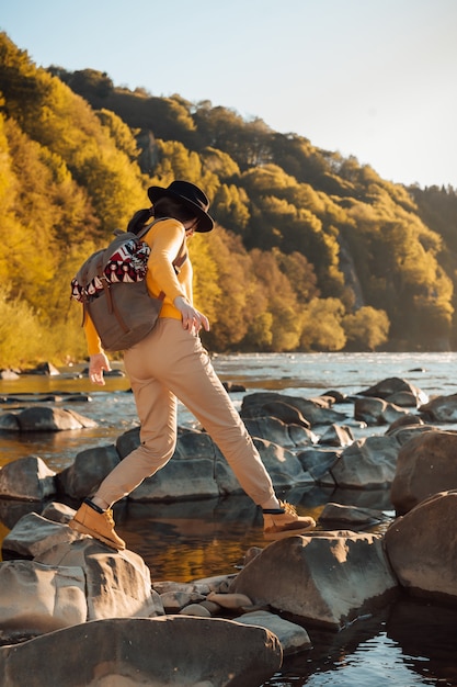 若い女性の旅行者は川の自然の背景にバックパックで立つ