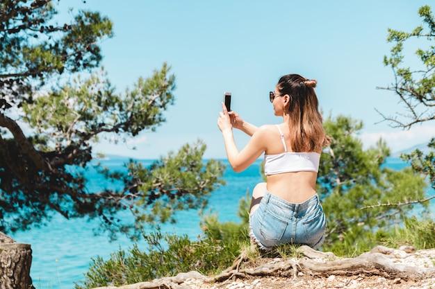 真昼の海の風景の写真を撮るサングラスの若い女性旅行者。青い海と松の木