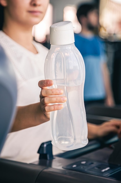 水のボトルを持ってジムのトレッドミルに乗る若い女性