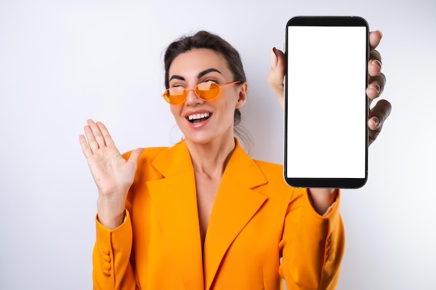 A young woman in trendy stylish glasses and a bright orange oversized jacket on a white background holds a phone with a blank white screen