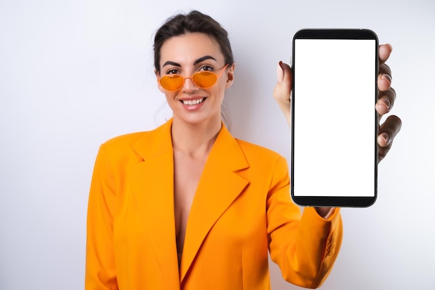 A young woman in trendy stylish glasses and a bright orange oversized jacket on a white background holds a phone with a blank white screen