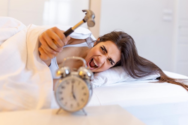 Young woman tries to break the alarm clock with hammer, destroy\
the clock. girl lying in bed turning off an alarm clock with hammer\
in the morning at 5am.